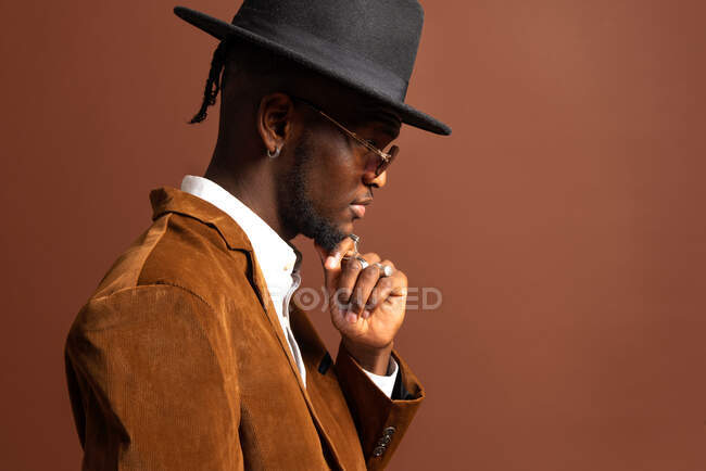 Вид сбоку на молодого афроамериканского мужчину в модной одежде и шляпе, смотрящего в сторону на коричневом фоне — стоковое фото