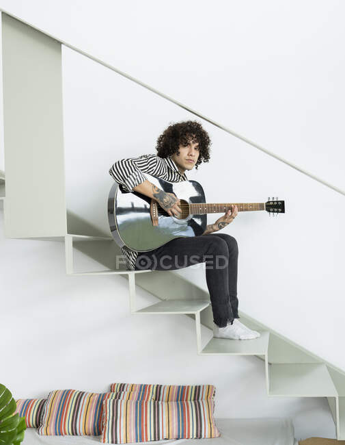 Side view of young hipster male musician playing acoustic guitar while sitting on staircase and looking at camera in flat — Stock Photo
