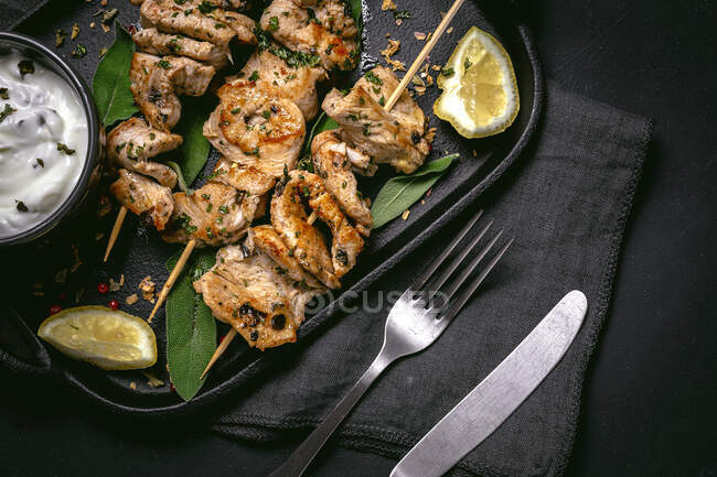 From above of appetizing freshly cooked meat on skewers served on tray on black table with bowl of sauce — Stock Photo