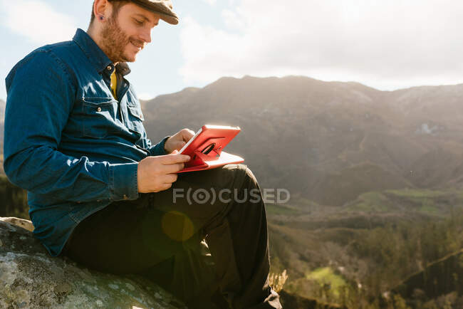 Seitenansicht des ruhigen männlichen Entdeckers mit Tablette, die auf einem Felsen sitzt und an sonnigen Tagen spektakulären Blick auf die Berge genießt — Stockfoto