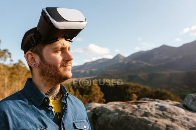 Viajante masculino curioso sentado na colina e experimentando a realidade virtual em óculos de proteção nas montanhas no dia ensolarado — Fotografia de Stock