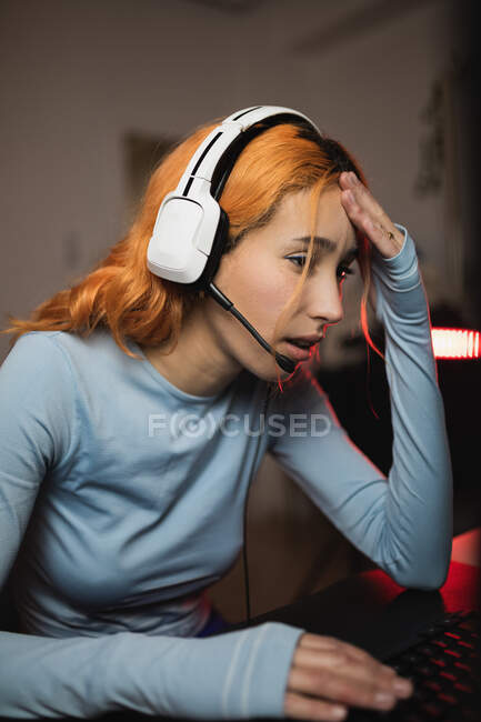 Вид сбоку обеспокоенной женщины в наушниках, играющей в видеоигру, сидя за столом с клавиатурой — стоковое фото