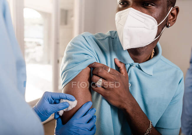 Cropped unrecognizable medical specialist in protective uniform and latex gloves disinfecting arm with cotton with alcohol to vaccinate male African American patient in clinic during coronavirus outbreak — Stock Photo