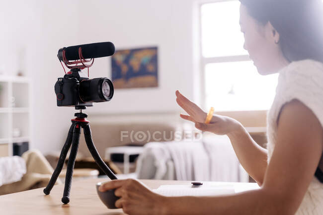 Vista lateral de la joven blogger étnica femenina con portátil sentado en la mesa con cámara de fotos en el trípode en la cocina - foto de stock