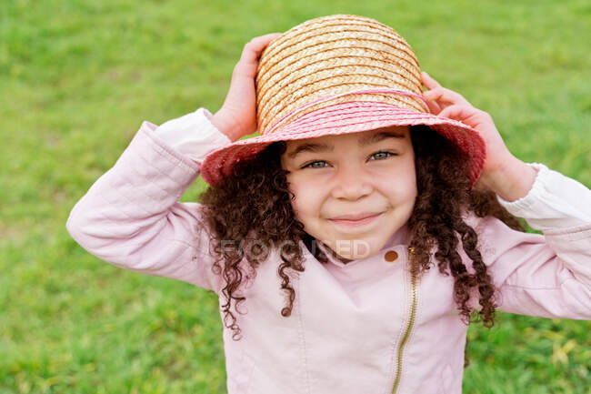 Fröhliches Kind in lässiger Kleidung mit lockigem Haar, das tagsüber auf dem grünen Rasen in die Kamera blickt — Stockfoto