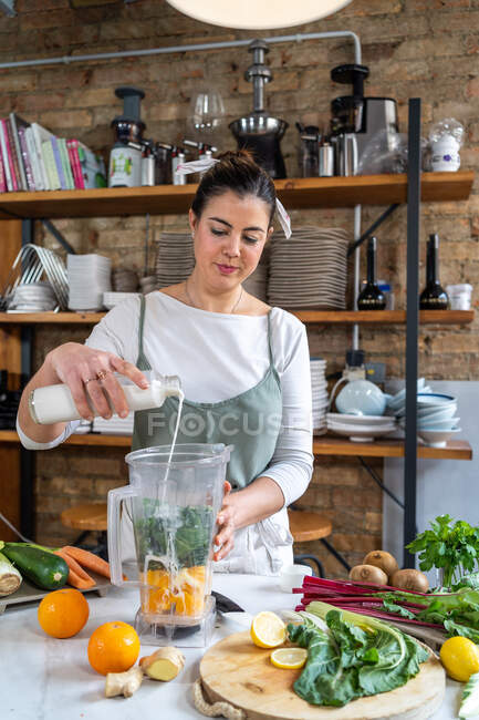 Femmina adulta versando latte vegetariano in ciotola con foglie di bietola e fette di arancia mentre prepara frullato a casa — Foto stock