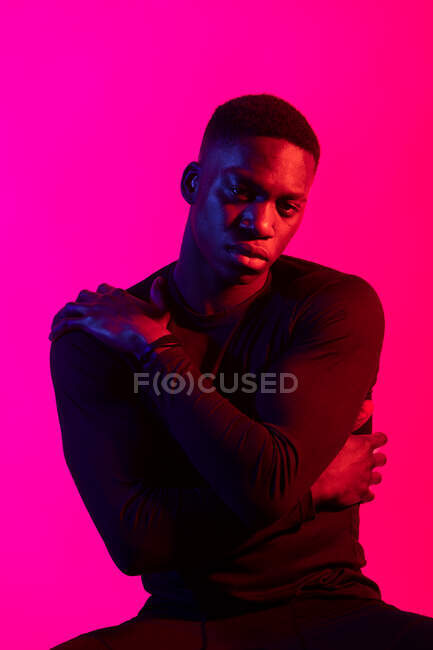 Homem negro jovem confiante em roupas esportivas escuras olhando para a câmera no fundo rosa néon no estúdio escuro — Fotografia de Stock