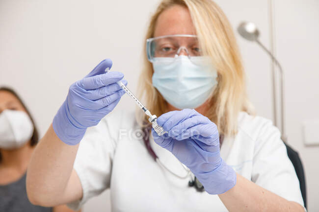 Female medic in protective face shield mask and latex gloves with vial of coronavirus vaccine and syringe showing to camera while standing in hospital room — Stock Photo