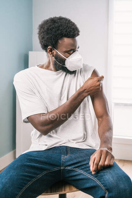 Paciente afroamericano que sostiene algodón con el brazo desinfectante de alcohol después del procedimiento de vacuna covid en la clínica durante el brote de coronavirus - foto de stock