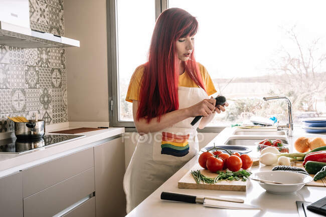 Giovane femmina in grembiule con simbolo LGBTQ peeling cetriolo fresco mentre prepara il pranzo in cucina casa luce — Foto stock