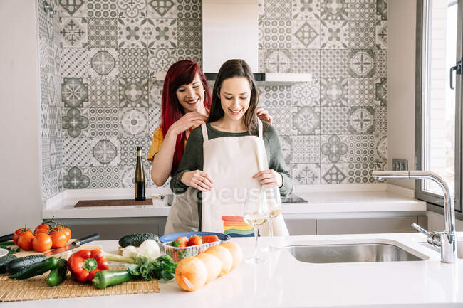 Smiling young woman putting apron on homosexual female beloved at table with assorted fresh vegetables in house — Stock Photo