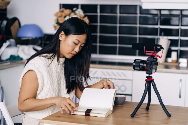 Молода етнічна жінка відеоблогер з блокнотом, що сидить за столом з фотоапаратом на тринозі на кухні — стокове фото