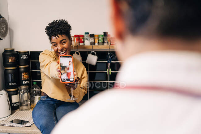 Fröhliche Afroamerikanerin zeigt Smartphone-Foto für unkenntlichen Freund beim Spaß in der heimischen Küche — Stockfoto