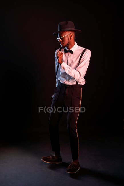 Junges männliches Model mit Hut und Hose steht tanzend vor schwarzem Hintergrund und schaut weg — Stockfoto