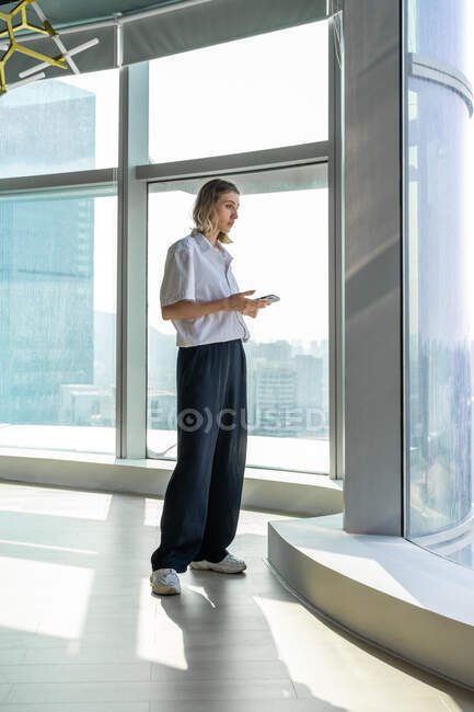 Einsame, emotionslose junge Frau steht in leerem Büro mit großem Fenster und surft auf dem Handy — Stockfoto