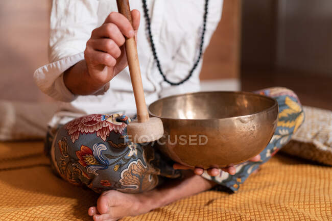 Crop unrecognizable Zen teacher with mallet playing Tibetan singing bowl while sitting with crossed legs on plaid — Stock Photo