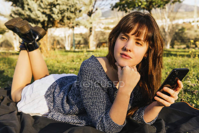 Vista lateral de la mujer despreocupada acostada en el prado a cuadros y usando el teléfono inteligente mientras disfruta de un picnic en el día soleado en primavera - foto de stock