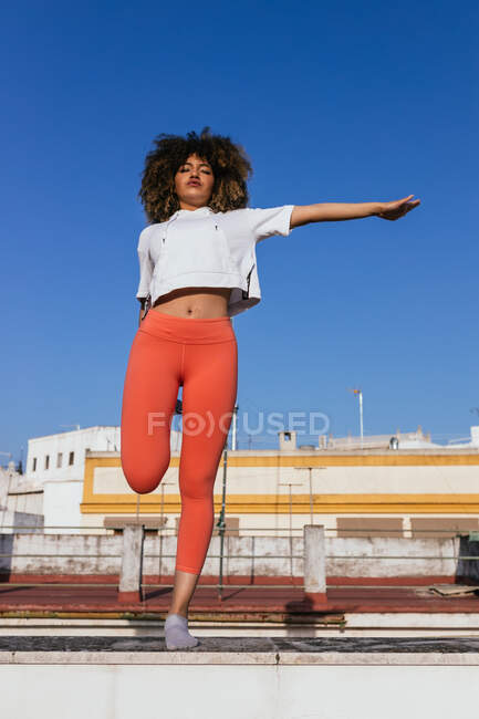Baixo ângulo de calma afro-americana fêmea em sportswear de pé em uma perna e com os olhos fechados enquanto pratica ioga no telhado no dia ensolarado — Fotografia de Stock