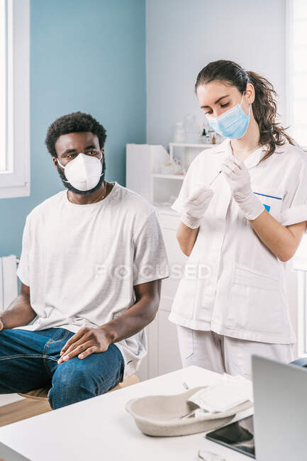 Female doctor in latex gloves and face shield filling in syringe from bottle with vaccine preparing to vaccinate unrecognizable African American man patient in clinic during coronavirus outbreak — Stock Photo