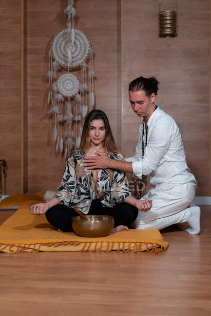 Professeur zen en vêtements blancs touchant la poitrine de la femme avec les jambes croisées pendant la pratique du yoga près du gong bol — Photo de stock