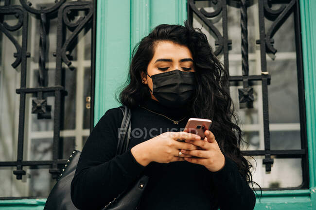 Bajo ángulo de graves más tamaño femenino en máscara protectora navegar por Internet en el teléfono inteligente, mientras que de pie en la calle cerca del edificio durante coronavirus - foto de stock