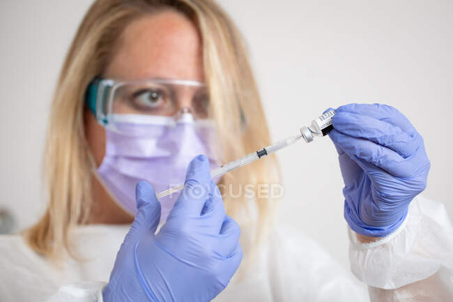 Médico femenino con mascarilla protectora y guantes de látex con vial de vacuna contra el coronavirus y jeringa que muestra a la cámara mientras está de pie en la habitación del hospital - foto de stock