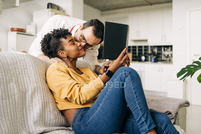 Side view of tender man kissing black woman sitting on sofa and reading book at weekend — Stock Photo