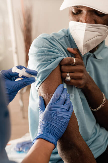 Médecin cultivé en uniforme de protection et gants en latex vaccinant le patient afro-américain masculin méconnaissable en clinique pendant l'épidémie de coronavirus — Photo de stock