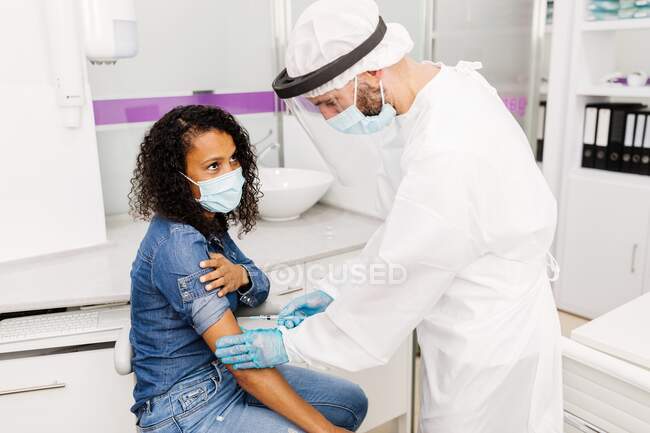Seitenansicht eines männlichen Facharztes in Schutzuniform, Latexhandschuhen und Gesichtsschutzimpfung einer afroamerikanischen Patientin in der Klinik während des Coronavirus-Ausbruchs — Stockfoto