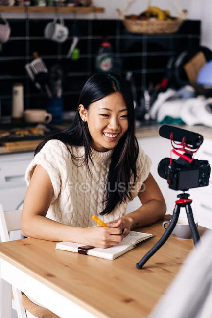 Молода етнічна весела жінка відеоблогер з блокнотом, що сидить за столом з фотоапаратом на тринозі на кухні — стокове фото