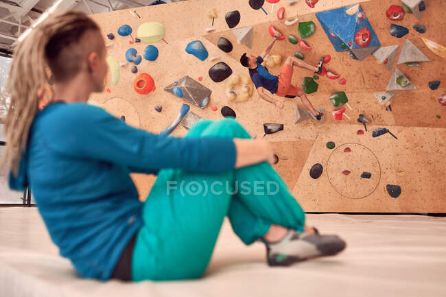 Vista lateral de la escaladora femenina observando fuerte macho escalada pared artificial en el club de bouldering moderno - foto de stock