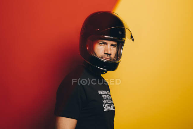 Vue latérale de l'homme adulte brutal confiant en casque de moto noir regardant la caméra tout en se tenant debout sur fond rouge et jaune coloré — Photo de stock