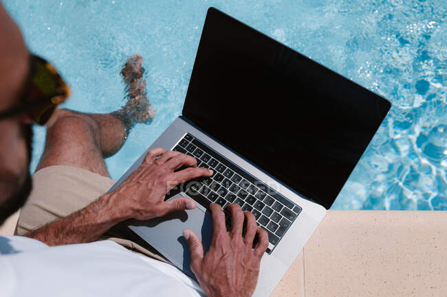 De cima vista traseira do freelancer masculino em óculos de sol sentado à beira da piscina e netbook de navegação enquanto trabalhava remotamente no projeto durante as férias de verão — Fotografia de Stock