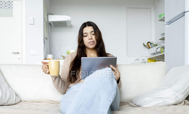 Молода жінка з планшетом і чашкою гарячого напою відпочиває з схрещеними ногами на дивані у вітальні — стокове фото