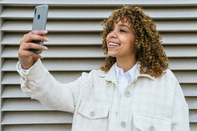 Mujer afroamericana optimista con peinado afro que toma autorretrato en un teléfono inteligente mientras está de pie contra la pared metálica en el área urbana de la ciudad - foto de stock