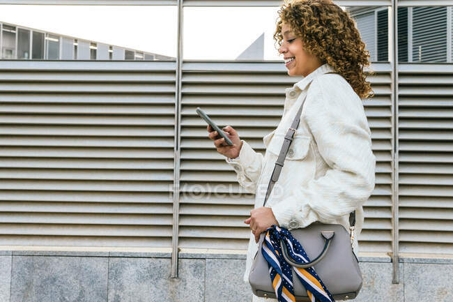 Side view of optimistic African American female with Afro hairstyle browsing on smartphone while standing against metal wall in urban area in city — Stock Photo