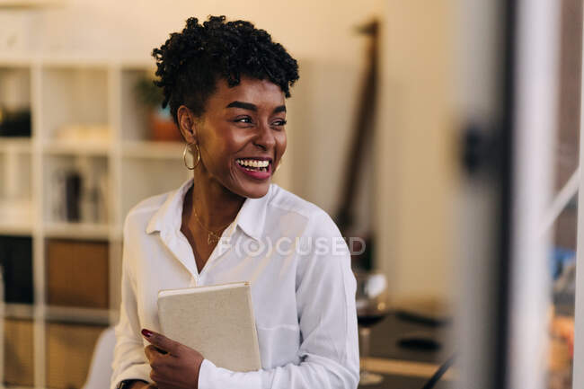 Fröhliche afroamerikanische Unternehmerin in weißem Hemd steht im Raum und hält Notizblock in der Hand, während sie von zu Hause aus arbeitet und wegschaut — Stockfoto