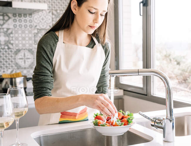 Colheita jovem fêmea em avental lavagem de morangos frescos sob fluido de água da torneira na cozinha da casa — Fotografia de Stock