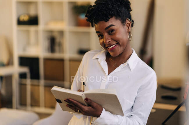 Cheerful African American female entrepreneur in white shirt standing in room and writing in notepad while working form home and looking at camera — Stock Photo