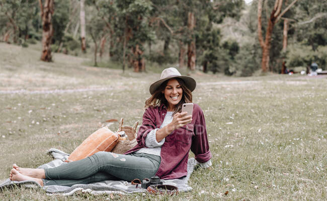 Весела жінка в капелюсі сидить на лузі в лісі і користується мобільним телефоном, насолоджуючись пікніком в Австралії. — стокове фото