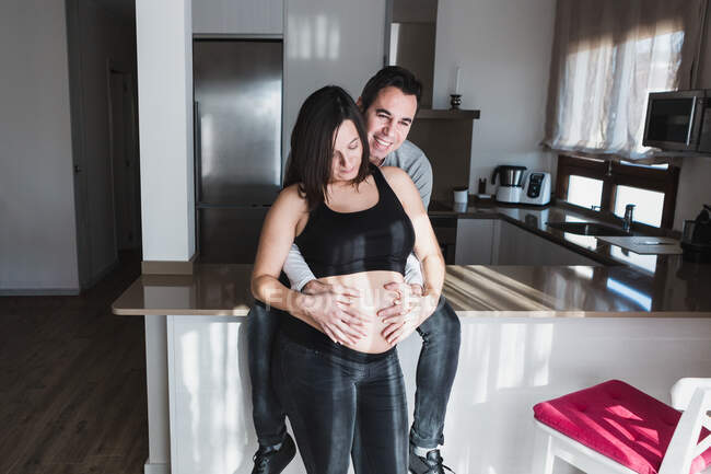 Homem encantado tocando barriga de mulher grávida em casa enquanto desfruta de fim de semana juntos — Fotografia de Stock