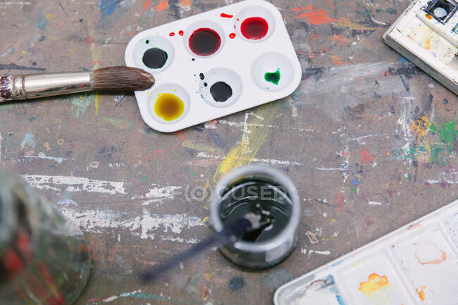Vista superior de pinceles y paletas de pintura dispuestas sobre mesa con tarro de agua en estudio de arte - foto de stock