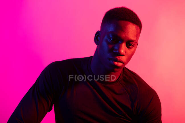 Уверенный молодой черный мужчина в темном спортивном наряде смотрит на камеру на неоновом розовом фоне в темной студии — стоковое фото