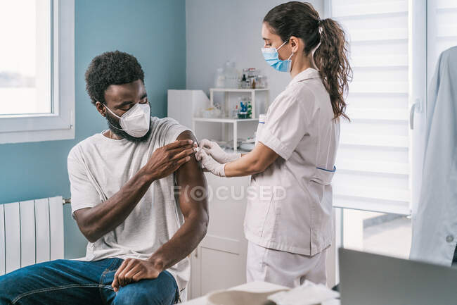 Especialista médica femenina en uniforme protector, guantes de látex y mascarilla facial vacunando a un paciente afroamericano en clínica durante el brote de coronavirus - foto de stock