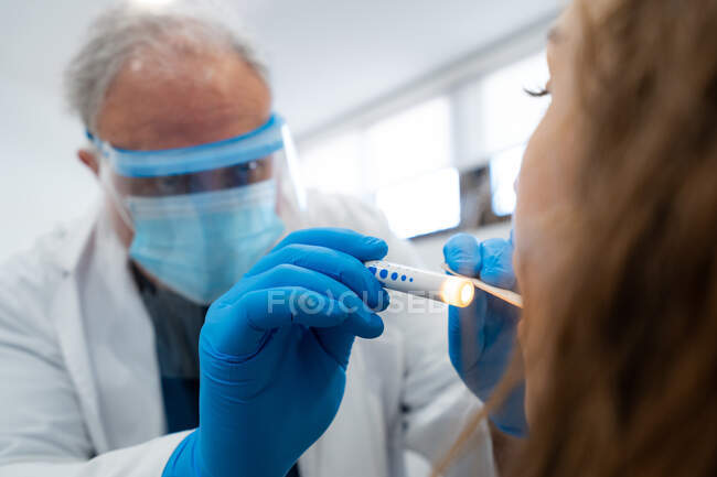 Otorrinolaringologista do sexo masculino em escudo protetor verificando a garganta do paciente com espátula estéril e lanterna elétrica no hospital — Fotografia de Stock