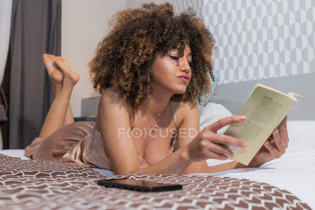 Etnia jovem atraente fêmea em roupa de noite com penteado afro e pernas cruzadas lendo livro didático enquanto deitado na cama — Fotografia de Stock