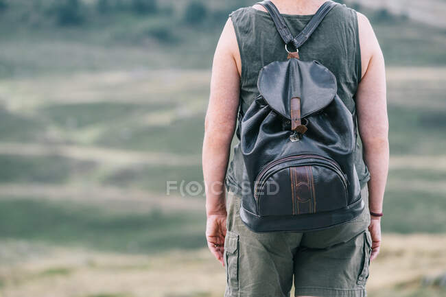 Vista posterior de una excursionista anónima con mochila de pie en la colina en un terreno montañoso y admirando el pintoresco paisaje - foto de stock