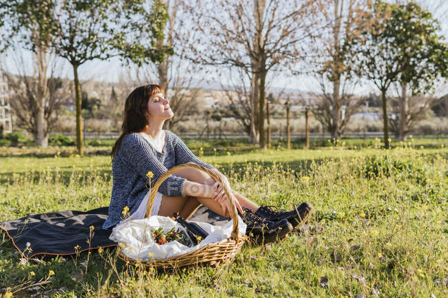 Vista lateral do conteúdo feminino sentado no cobertor perto da cesta e desfrutando do sol durante o piquenique no prado na primavera — Fotografia de Stock