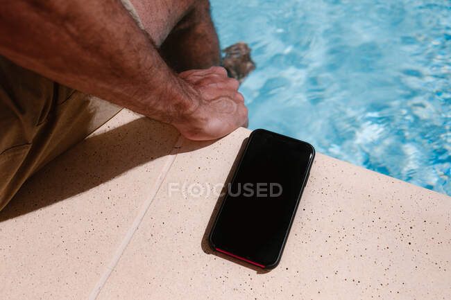 Зверху врожаю невпізнаваний чоловічий фрілансер сидить біля басейну біля смартфона з чорним екраном під час віддаленої роботи влітку — стокове фото