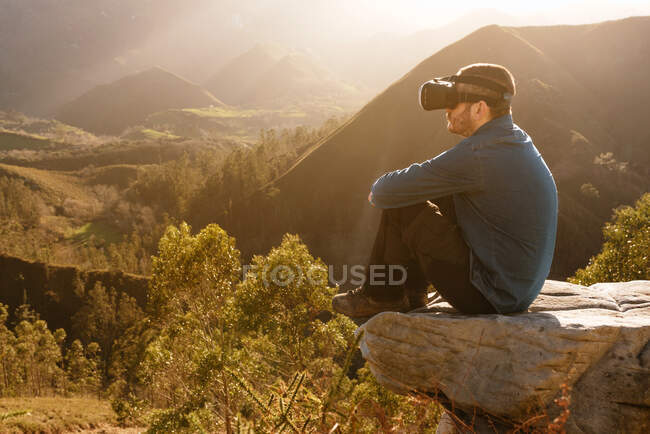 Вид збоку чоловічого мандрівника в окулярах VR, що взаємодіють з віртуальною реальністю, сидячи на пагорбі в гірській місцевості на заході сонця — стокове фото
